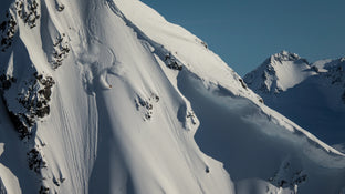 A skier descends a steep, snow-covered mountain slope on the Icelantic Skis Nomad 112, carving tracks in the fresh powder under clear blue skies with rugged snowy peaks rising in the background.