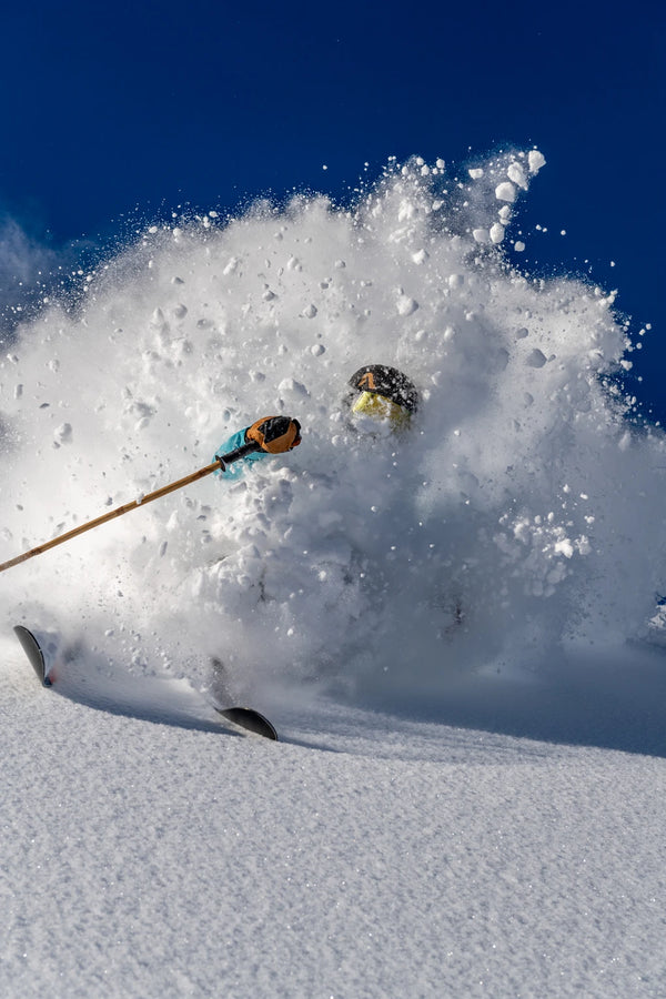 A skier charges through fresh powder on Icelantic Skis’ Nomad 112, nearly disappearing in a cloud of snow with only their ski poles, goggles, and the tips of the Nomad 112s visible against a backdrop of clear blue sky.