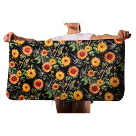 A person holds the Icelantic Skis Sunflower Travel Towel, featuring an ultra-soft rectangular design adorned with vibrant sunflowers and greenery on a black background, with only their arms and torso visible.