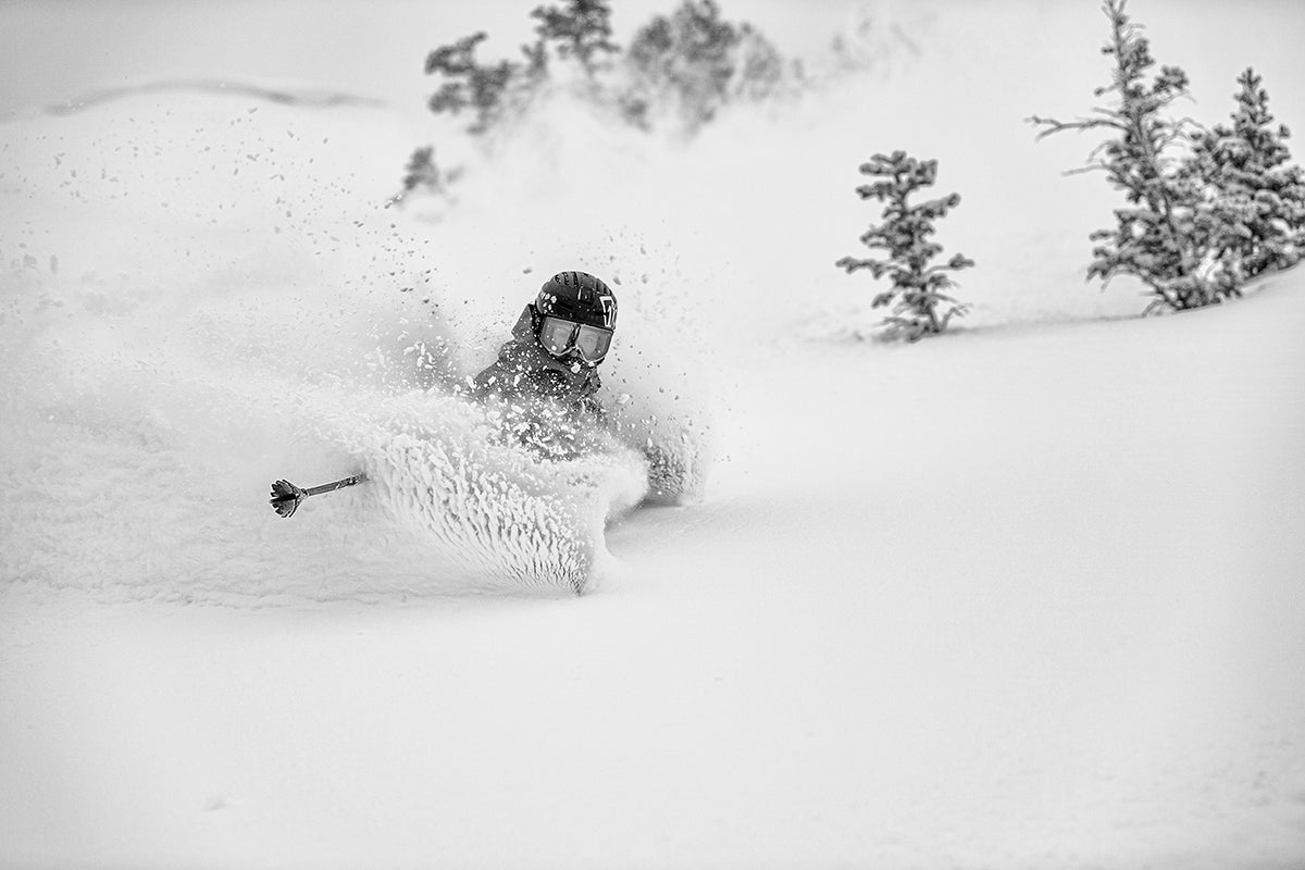 A skier in goggles and a helmet carves through deep powder snow, creating a spray, with small snow-covered trees visible in the background. The image is in black and white.