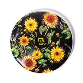 A black Sunflower Frisbee by Icelantic Skis, adorned with vibrant yellow sunflowers and green leaves, featuring a yellow shield-shaped logo with the white letters “T K” at the center, reminiscent of Travis Parr artwork.