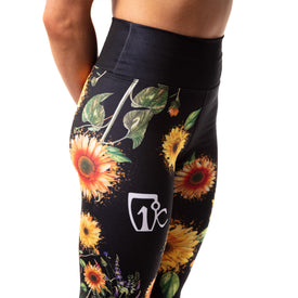 A person is wearing the Sunflower Nia Pro Legging by Icelantic Skis, which features a black base with a sunflower and leaf pattern, a white logo on the thigh, and a high-waisted waistband for added support, photographed from the side against a white background.