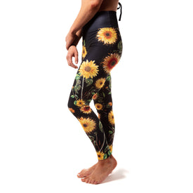 A person stands barefoot in Icelantic Skis’ Sunflower Nia Pro Legging, featuring a vibrant sunflower print on black and a high-waisted waistband, shown from the side against a white background.