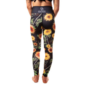 A person stands barefoot, facing away, wearing the Sunflower Nia Pro Legging from Icelantic Skis. These high-waisted black leggings feature a vibrant sunflower and leaf pattern, with an Icelantic logo and branding on the waistband. Crafted from advanced Hyper Wiking WIKMAX Fabric, the leggings accentuate only their lower back and legs.