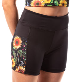 A person wears the Icelantic Skis Sunflower Pocket Short, stylish black women’s summer shorts made from recycled fabric and featuring a sunflower and butterfly pattern on the side pocket. Only the lower torso and upper legs are visible.