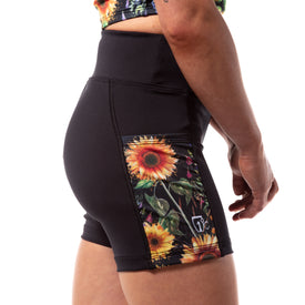 A person is shown from the waist down and viewed from the side against a white background, wearing the Icelantic Skis Sunflower Pocket Short—black women’s summer shorts featuring a vibrant sunflower and floral patterned side pocket, crafted from recycled fabric.