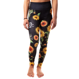 A person stands barefoot against a white background, wearing Icelantic Skis’ Sunflower Nia Pro Legging—high-waisted black leggings adorned with vibrant yellow sunflower prints. These stylish and comfortable leggings are crafted from Hyper Wiking WIKMAX Fabric. The upper body is partially visible with arms relaxed at their sides.