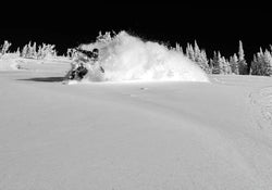 A skier on Icelantic Skis’ Nomad 112 Whiteout carves through deep powder, sending up a burst of snow spray as snow-covered trees and a dark sky form the dramatic backdrop.