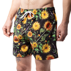 A person wearing Icelantic Skis Sunflower Swim Trunks, featuring a vibrant sunflower and green leaf pattern on a black background. These eco-friendly swim trunks offer both style and comfort with their elastic waistband and white drawstring, making them the perfect choice for your next beach outing.