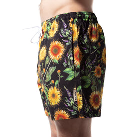A person shown from the side against a white background wears Icelantic Skis Sunflower Swim Trunks, featuring a black base with a vibrant yellow sunflower and green leaf pattern.
