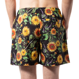 A person is seen from the back against a white background, wearing Icelantic Skis’ eco-friendly Sunflower Swim Trunks featuring a bold sunflower and green leaf pattern—perfect for making a statement by the water.