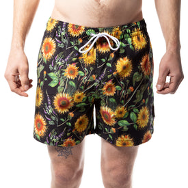 A person wearing Icelantic Skis Sunflower Swim Trunks, featuring a bright sunflower and green leaf pattern, stands against a white background.