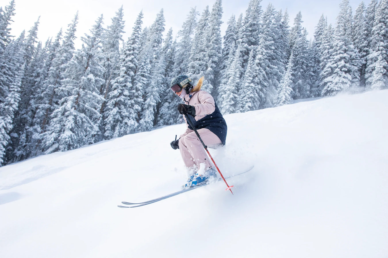 A skier wearing a pink ski suit and helmet descends a snowy slope on Icelantic Skis’ Maiden 102, surrounded by snow-covered evergreen trees on a bright winter day.