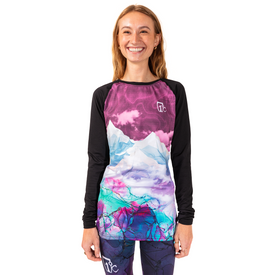 A smiling person wearing the Icelantic 23/24 Womens WikMax Baselayer Top from the Riveter Collection, which features vibrant mountain and sky designs, stands against a white background.