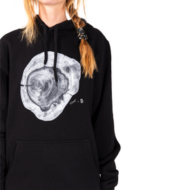 A woman wears a black Icelantic Yew Lineage Hoodie from the Jack Ludlam Backcountry Collection, featuring a bold white tree ring graphic on the front. She stands against a white background, with only her upper body and part of her face visible.
