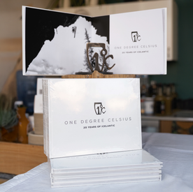 A stack of One Degree Celsius: 20 Years of Icelantic photo books from Icelantic Skis sits on a table, with one book open to a black-and-white nature photograph.