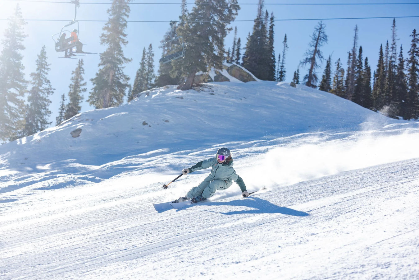 A skier on Icelantic Skis’ Maiden 94, dressed in a light blue outfit, carves down a sunny, snowy slope with trees and a ski lift in the background.