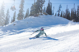 A skier on Icelantic Skis’ Maiden 94, dressed in a light blue outfit, carves down a sunny, snowy slope with trees and a ski lift in the background.