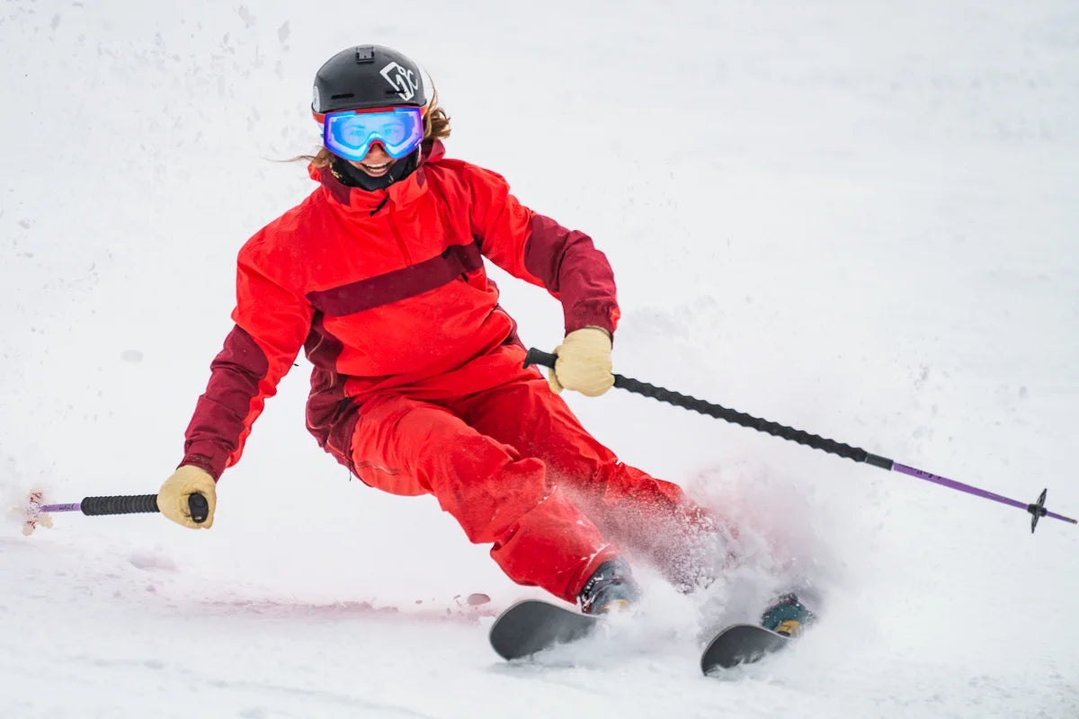 Clad in a red outfit and blue goggles, a skier expertly carves a sharp turn through the snow—sending up a spray of powder—as they grip their ski poles and wear a helmet. They’re riding on the Maiden 94 by Icelantic Skis.