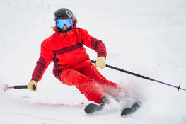 Clad in a red outfit and blue goggles, a skier expertly carves a sharp turn through the snow—sending up a spray of powder—as they grip their ski poles and wear a helmet. They’re riding on the Maiden 94 by Icelantic Skis.