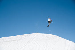 A skier catches air on Icelantic Skis’ Nomad 100, soaring above a snowy slope with snow swirling behind against a clear blue sky.