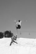 A skier catches air above a rail on a snowy slope, showcasing the Icelantic Skis Nomad 94 Whiteout, with ski poles extended and chairlifts in the background under a clear sky.