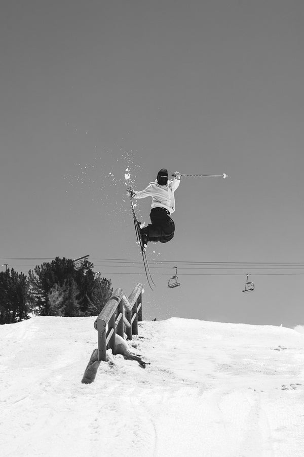 A skier catches air above a rail on a snowy slope, showcasing the Icelantic Skis Nomad 94 Whiteout, with ski poles extended and chairlifts in the background under a clear sky.