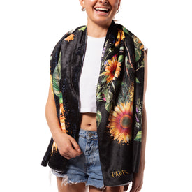 A smiling person in a sleeveless white crop top and denim shorts shows off the Sunflower Towel by Icelantic Skis—a black towel featuring large, colorful sunflower artwork inspired by Travis Parr, with "PATR" embroidered in yellow on the bottom corner.