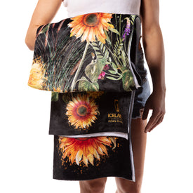 A person in a white tank top and denim shorts holds the Sunflower Towel from Icelantic Skis—a folded microfiber towel featuring a vibrant sunflower design inspired by Travis Parr artwork—against a black background. This quick-dry towel combines eye-catching style with everyday practicality.