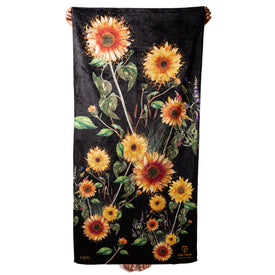 A person holds up the Icelantic Skis Sunflower Towel, a rectangular black microfiber towel featuring a vibrant design of yellow sunflowers, greenery, and scattered small purple flowers. Only the person’s hands and feet are visible. This quick dry towel beautifully showcases Travis Parr’s artwork, adding an artistic flair to everyday essentials.