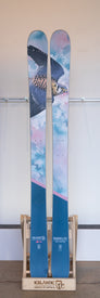A pair of blue Icelantic Demo - 23/24 Maiden Lite skis—an ultra-light freeride model—stand upright in a wooden ski rack bearing the Icelantic brand against a light background, each featuring detailed artwork of a flying bird and clouds.