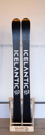 A pair of Icelantic Demo - 23/24 Maiden Lite skis in black with white lettering stand in a wooden ski holder labeled ICELANTIC, embodying the spirit of an ultra-lite freeride ski crafted for adventure, set against a plain background.