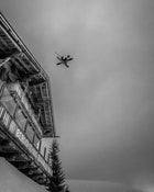 A skier launches off the roof of a rustic mountain lodge, soaring through the air on Icelantic Skis’ Nomad 112 Whiteout, captured in striking black and white against a backdrop of moody clouds.