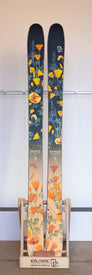 A pair of Icelantic Demo - 23/24 Nia Pro 105 skis with a floral design featuring orange and yellow flowers rests on a wooden stand labeled "Icelantic return to nature," highlighting the Reflective Rocker profile for enhanced performance.