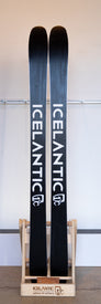 A pair of 23/24 Icelantic Nia Pro 105 skis in black, standing upright in a wooden holder, featuring the “ICELANTIC” brand name printed vertically in white lettering on each ski and highlighting their signature Reflective Rocker profile.