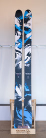 A pair of Demo - 23/24 Saba Pro 107 skis by Icelantic, featuring a blue, black, and white color scheme, stand upright in a wooden rack labeled "Icelantic Return to Nature" against a plain background, highlighting the signature Icelantic Reflective Rocker design.