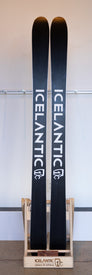 A pair of black Demo - 23/24 Saba Pro 107 skis by Icelantic stand upright in a wooden rack, with the Icelantic brand name and logo visible on both the skis and the rack, proudly showcasing their Reflective Rocker design.
