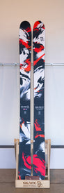 A pair of Icelantic Demo - 23/24 Saba Pro 117 skis featuring a red, black, and white abstract design stands upright on a wooden display rack against a plain wall, highlighting their innovative Reflective Rocker profile.