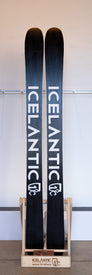 A pair of Demo - 23/24 Saba Pro 117 skis from Icelantic, featuring a black design with white lettering and the signature Reflective Rocker construction, stand upright on a wooden ski rack labeled “ICELANTIC return to nature” against a plain indoor wall.