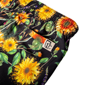 Close-up of Icelantic Skis’ Sunflower Swim Trunks in black, showcasing a vibrant sunflower and green leaf pattern. These eco-friendly swim trunks feature an elastic waistband, side pocket, and a small beige logo tag sewn onto the pocket—perfect for summer days.