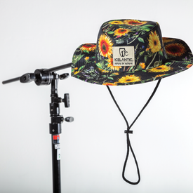 The Icelantic Skis Sunflower Boonie Hat, designed for sun protection, hangs from a black stand against a white background. This hat features bright yellow and orange floral patterns, a chin strap, and a patch that reads "ICELANTIC Nature is Culture," along with artwork by Travis Parr.