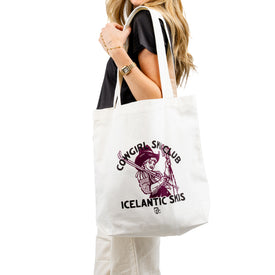A woman carries the Cowgirl Ski Club Tote by Icelantic, a sturdy and spacious cotton canvas bag featuring a purple graphic of a cowgirl holding skis and the text “Cowgirl Ski Club Icelantic Skis.”.