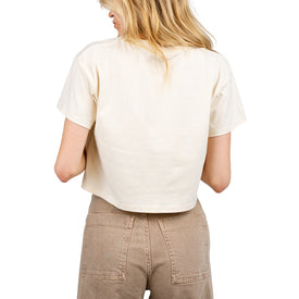 A person with blonde hair, seen from the back, wears the Icelantic Cowgirl Ski Club Cropped Tee in a relaxed fit cream style, paired with high-waisted light brown pants. Set against a white background, the look captures the effortless vibe of the Cowgirl Ski Club.