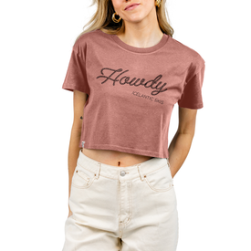A woman wears the Icelantic Howdy Cropped Tee in Hazy Pink—crafted from 100% combed cotton and featuring bold "Howdy" text—paired with white pants and gold hoop earrings. With one hand in her pocket, she stands against a white background, effortlessly evoking modern cowboy culture.