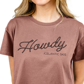 A woman is seen from the shoulders up wearing the Howdy Cropped Tee in Hazy Pink by Icelantic, featuring a rope-style "Howdy" script inspired by cowboy culture and "ICELANTIC SKIS" in smaller text beneath. This stylish women’s top is made from 100% combed cotton for a soft, comfortable fit.