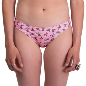 A person stands against a white background wearing the Icelantic Skier MTN Bikini Bottom in pink, featuring colorful yoga-inspired prints and a recycled swimwear look. Their hands, decorated with rings, hang relaxed at their sides.
