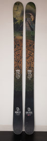A pair of Icelantic 20/21 Nomad 115 Used Demo skis, showcasing Aztec-inspired graphics with a pyramid and jungle motif, lean upright against a white wall—an excellent choice for anyone seeking a big mountain freeride ski that combines performance with standout style.