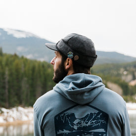 A person wearing an Icelantic Stormy Kromer Cap and a blue hoodie stands outdoors, facing away toward a forest and mountains, with snow visible on the ground.
