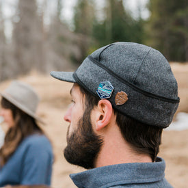 A man stands outdoors, facing away, wearing an Icelantic Stormy Kromer Cap in gray wool adorned with two pins on the side. In the blurred background, a woman in a hat is visible among trees and dry grass.