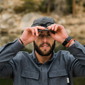 A bearded person in a gray button-up shirt wears the Icelantic Stormy Kromer Cap as they adjust their sunglasses outdoors, with a blurred natural background behind them.